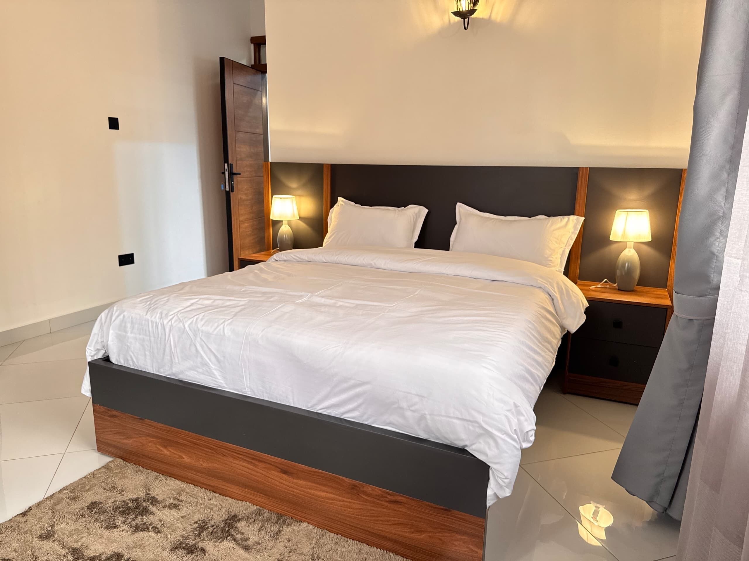 Una cama bien hecha con estructura de madera y ropa blanca, flanqueada por dos mesitas de noche con lámparas, en una habitación de hotel moderna.