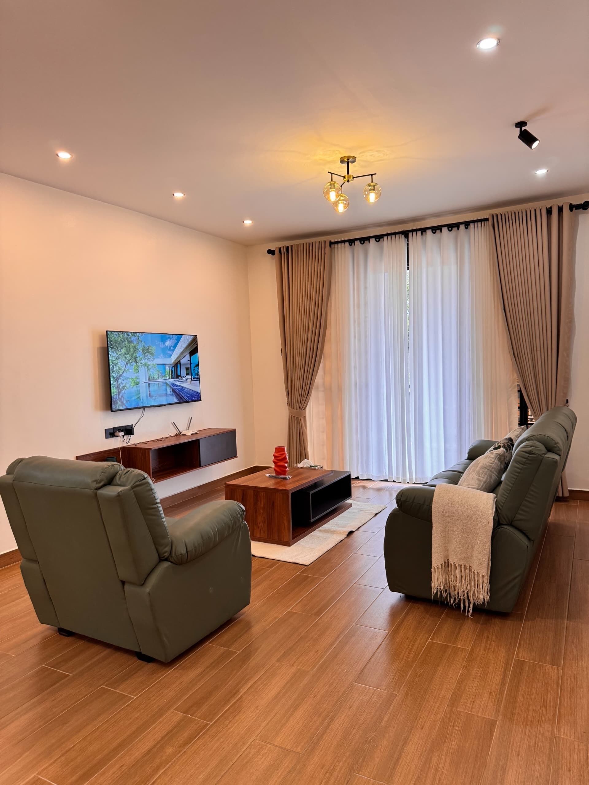 A modern living room with a flat-screen TV, a wooden coffee table, two green sofas, and a large window with curtains.