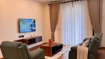 A modern living room with a flat-screen TV, a wooden coffee table, two green sofas, and a large window with curtains.