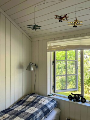 Una habitación con cama, ventana con vista a follaje y maquetas de aviones colgando del techo.
