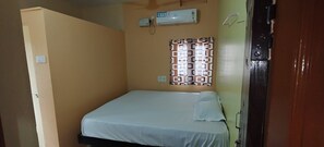 A small, single-bed room with a plain bed, a wall-mounted air conditioner, and a patterned window blind.