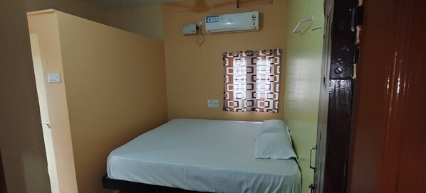 A small, single-bed room with a plain bed, a wall-mounted air conditioner, and a patterned window blind.
