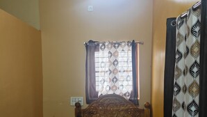 A single bed with a wooden headboard, a ceiling fan, a window with curtains, and a wall-mounted light fixture. - Gorgeous 6-bedroom house with AC in charming Sanapur (Sanapur)