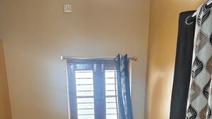 A single bed with a wooden frame and a mattress, a window with blue curtains, and a wall-mounted curtain. - Gorgeous 6-bedroom house with AC in charming Sanapur (Sanapur)