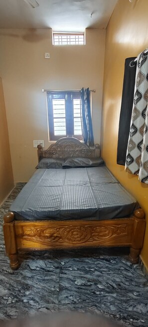 A single bed with a wooden frame and a mattress, a window with blue curtains, and a wall-mounted curtain. - Gorgeous 6-bedroom house with AC in charming Sanapur (Sanapur)
