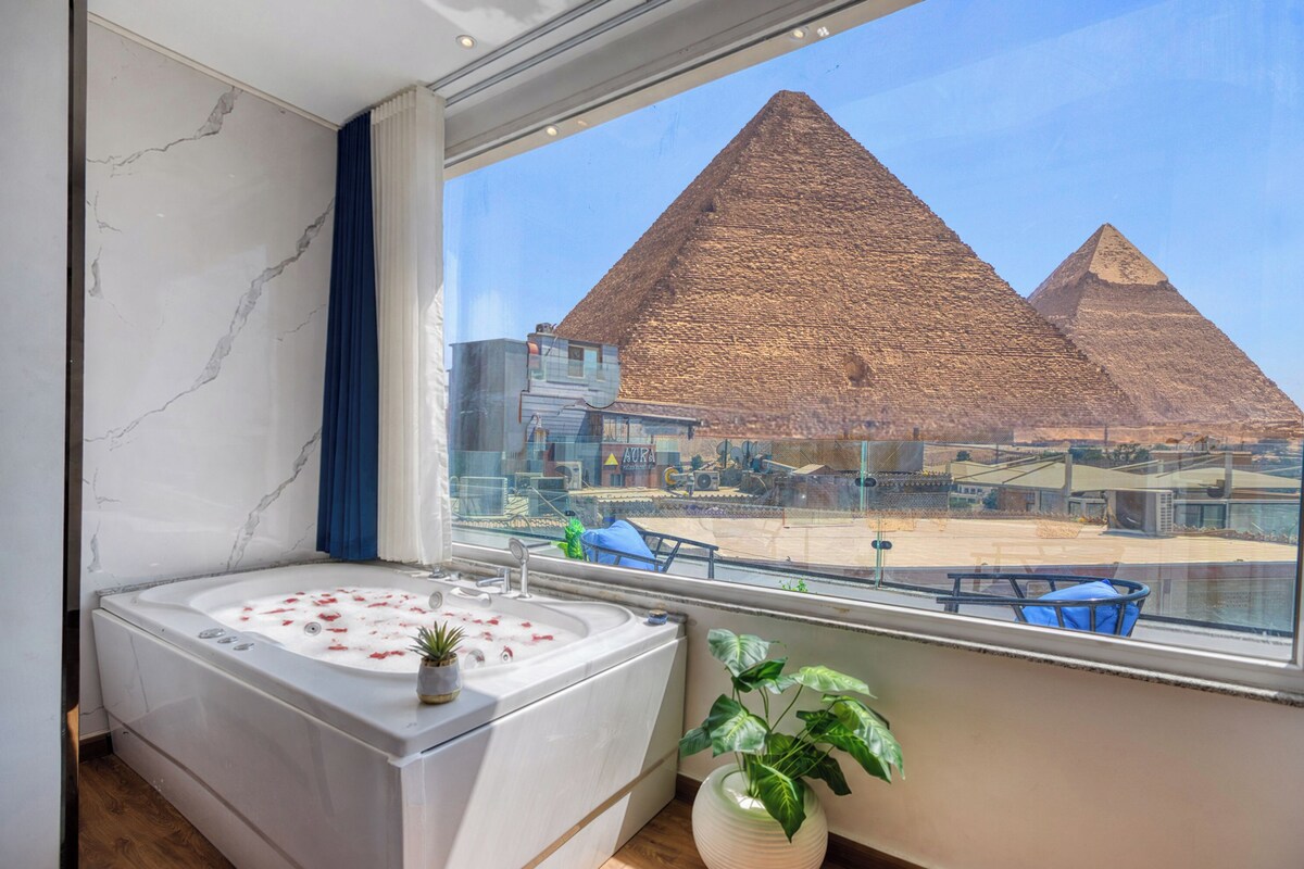 A modern bathroom with a large bathtub, a potted plant, and a view of the pyramids.