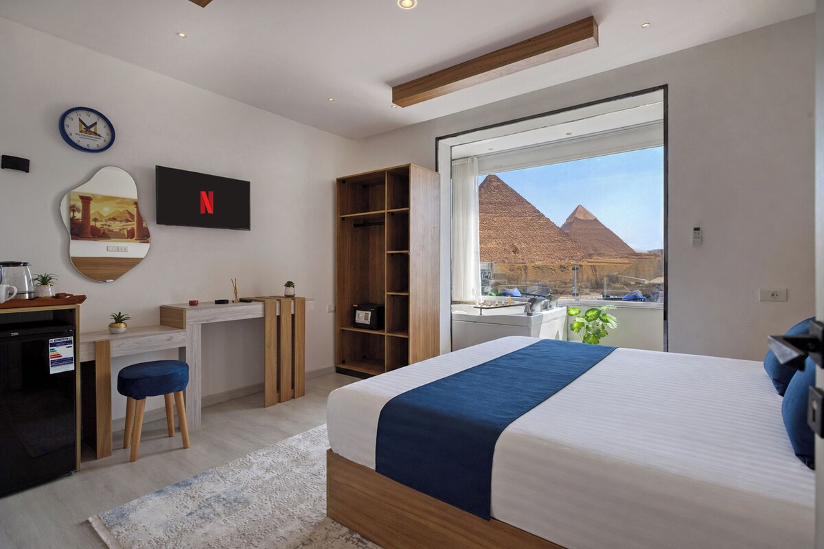 A modern bedroom with a bed, a desk, a wardrobe, and a large window with a view of pyramids.