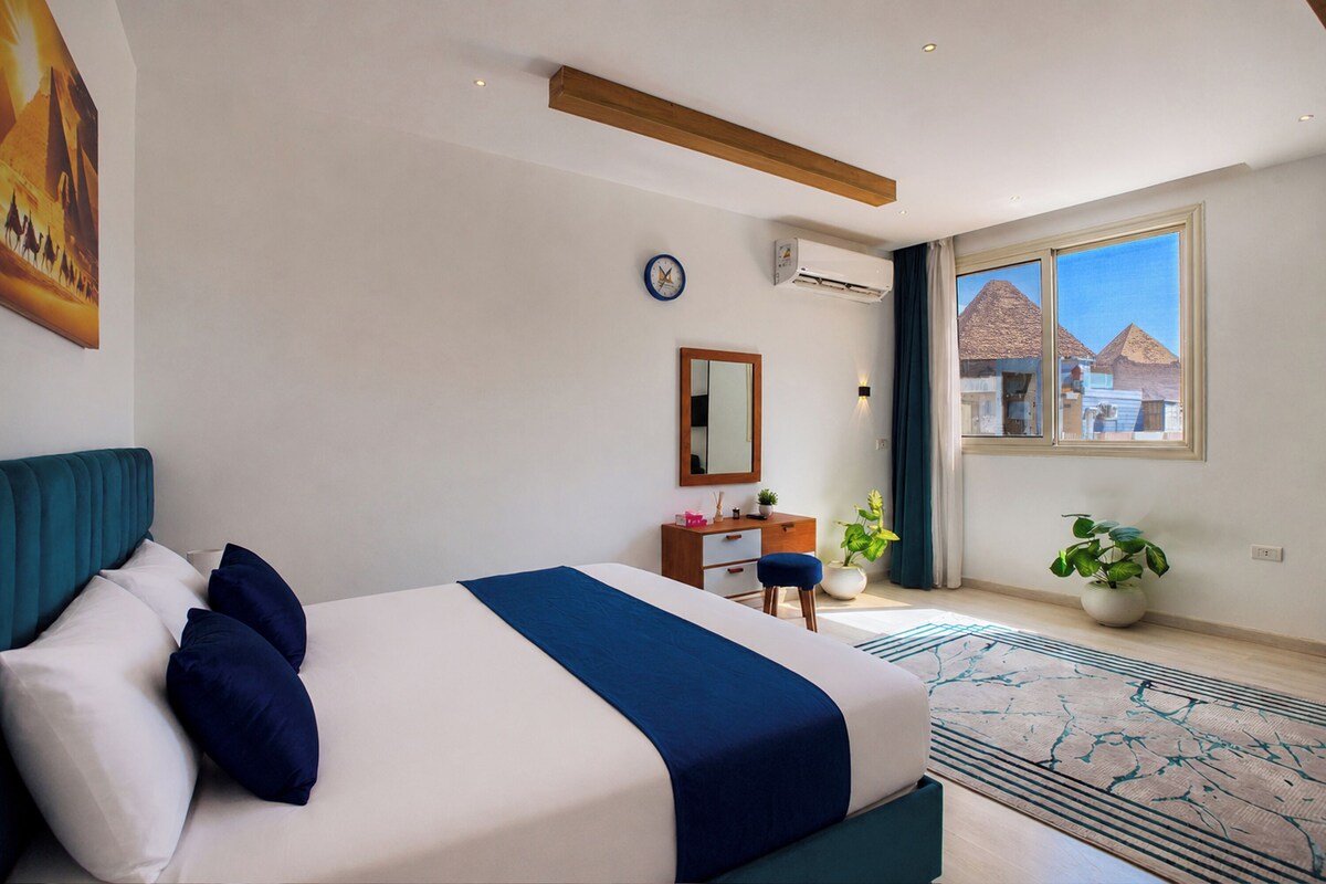 A hotel room with a bed, a dresser, a mirror, a clock, an air conditioning unit, a window with a view of pyramids, and a potted plant.