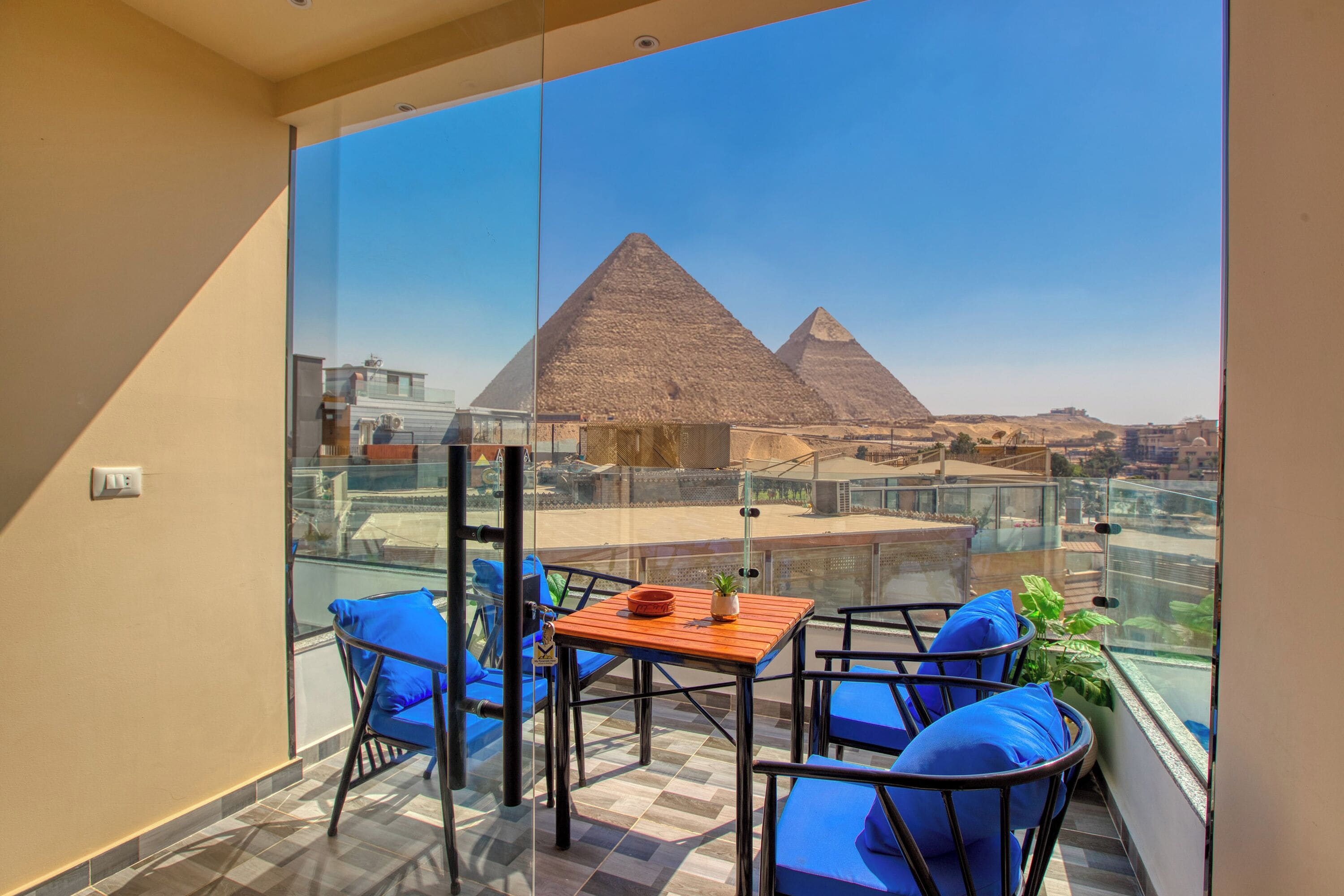 A balcony with a glass partition, blue cushioned chairs, a wooden table, and a view of the pyramids.
