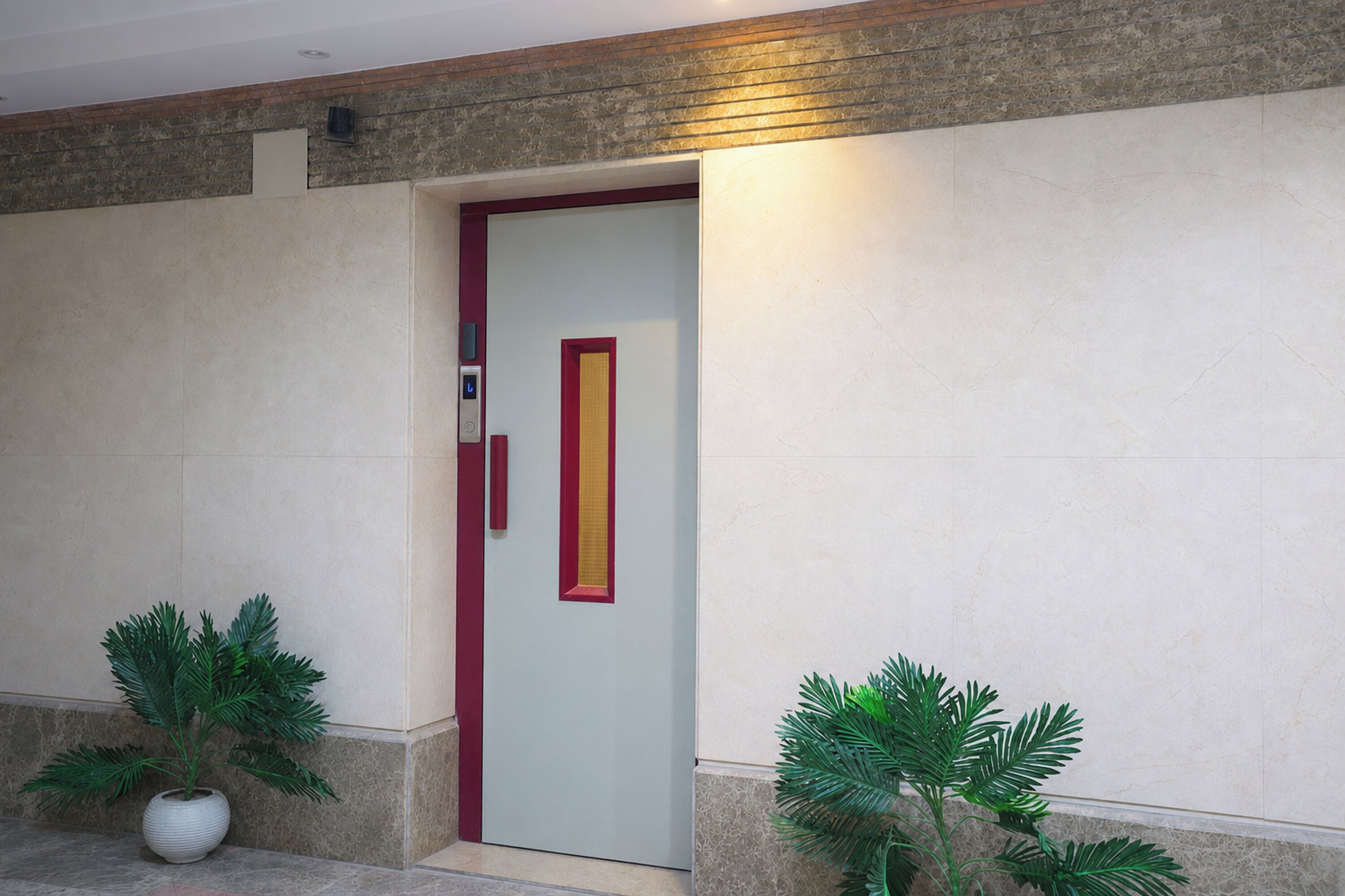 Entrada de hotel con una puerta de color claro, un marco rojo y una franja vertical. Hay dos macetas a cada lado de la puerta.