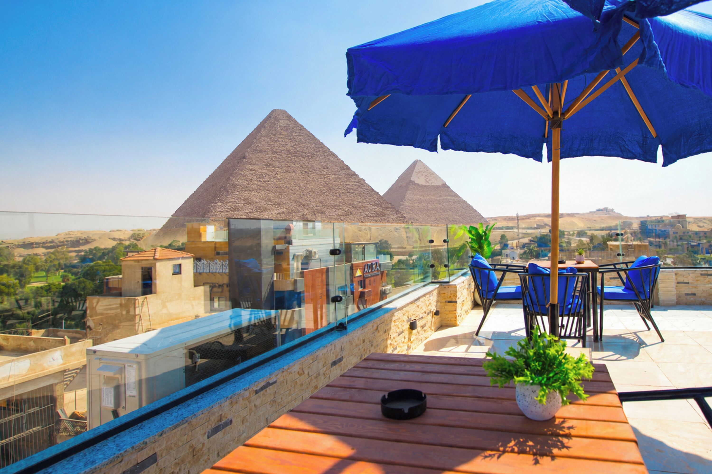 Une terrasse sur le toit, dotée d’une balustrade en verre offrant une vue sur les pyramides, une table avec des chaises et un pot de fleurs.