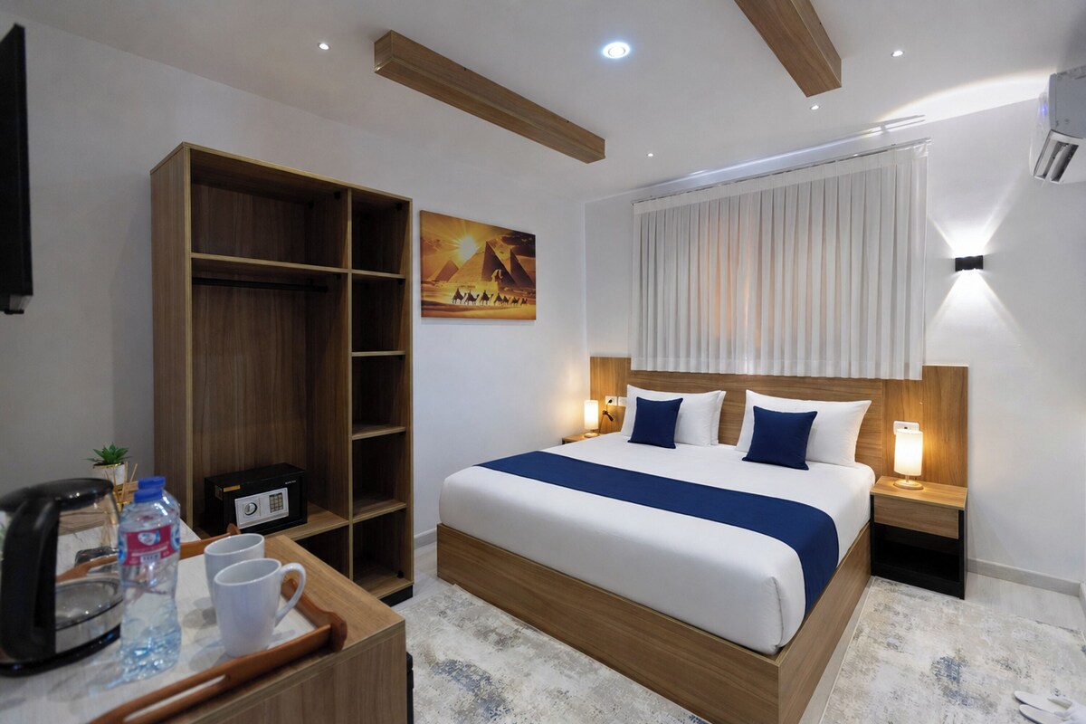 A hotel room with a bed, a wooden wardrobe, a desk with a coffee maker, and a painting of pyramids.