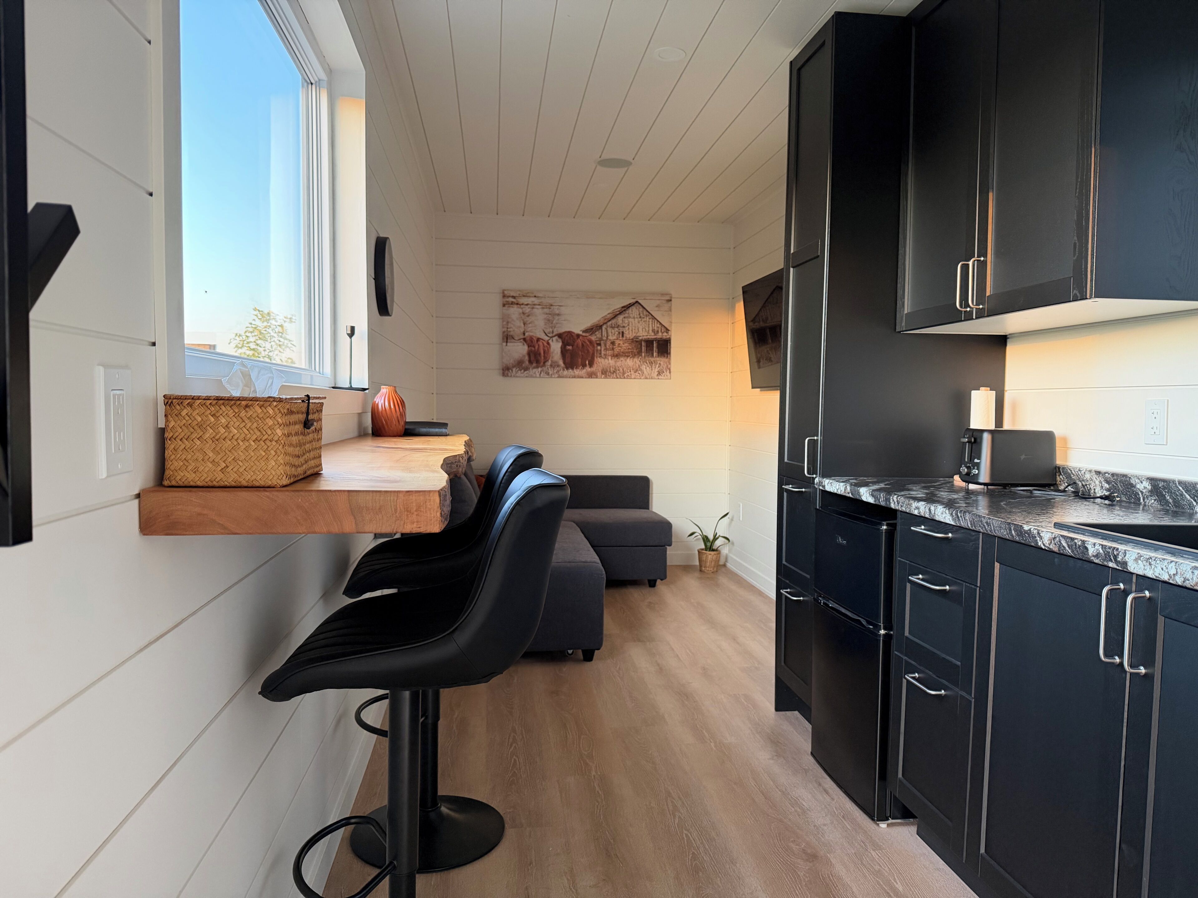 A modern kitchen with a built-in bar area, dark cabinets, and a large window.