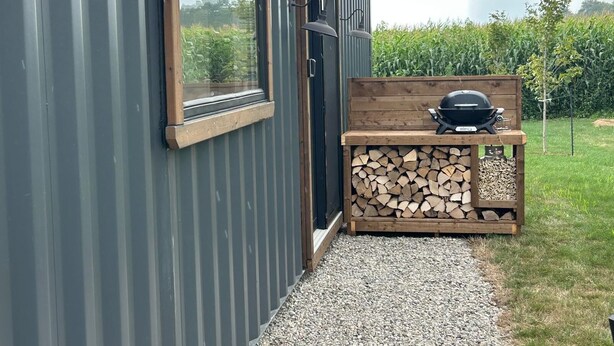 A modern building with a metal exterior, a wooden bench with a barbecue grill, and a stack of firewood.