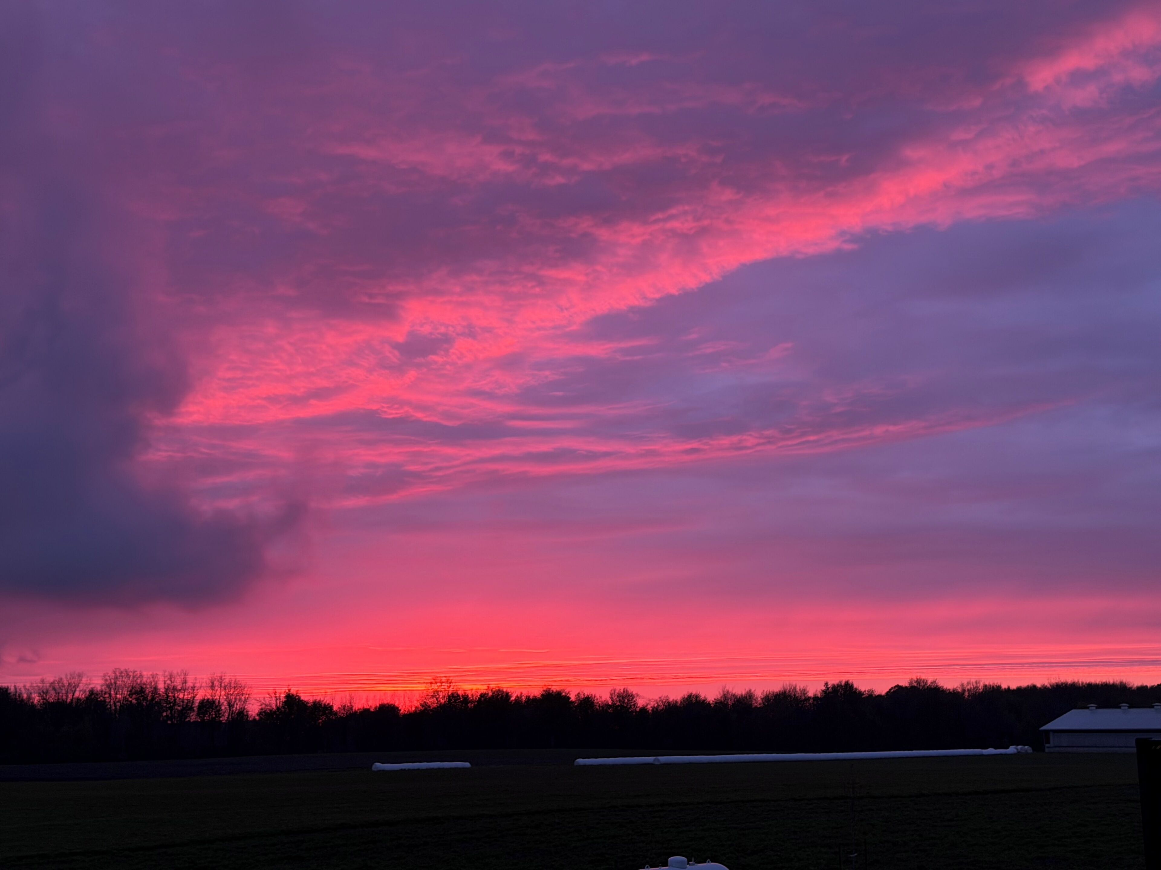 A rural landscape with a field, a building, and a sunset sky.