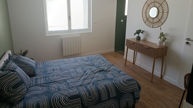 Une chambre à coucher comprenant un lit, une fenêtre, un radiateur, une console en bois avec un miroir et une porte.