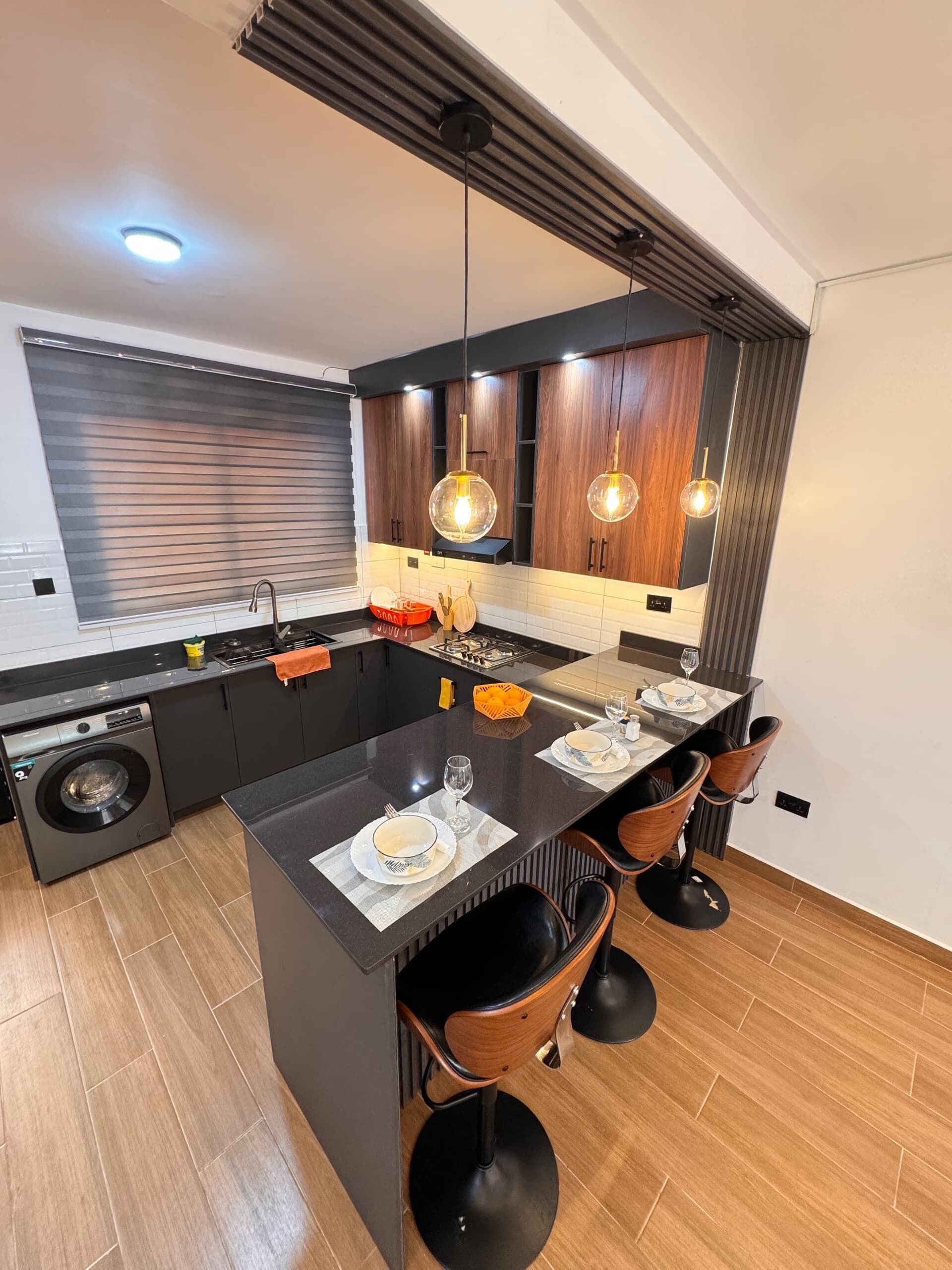 A modern kitchen with a central island, wooden cabinets, pendant lighting, and a built-in washing machine.