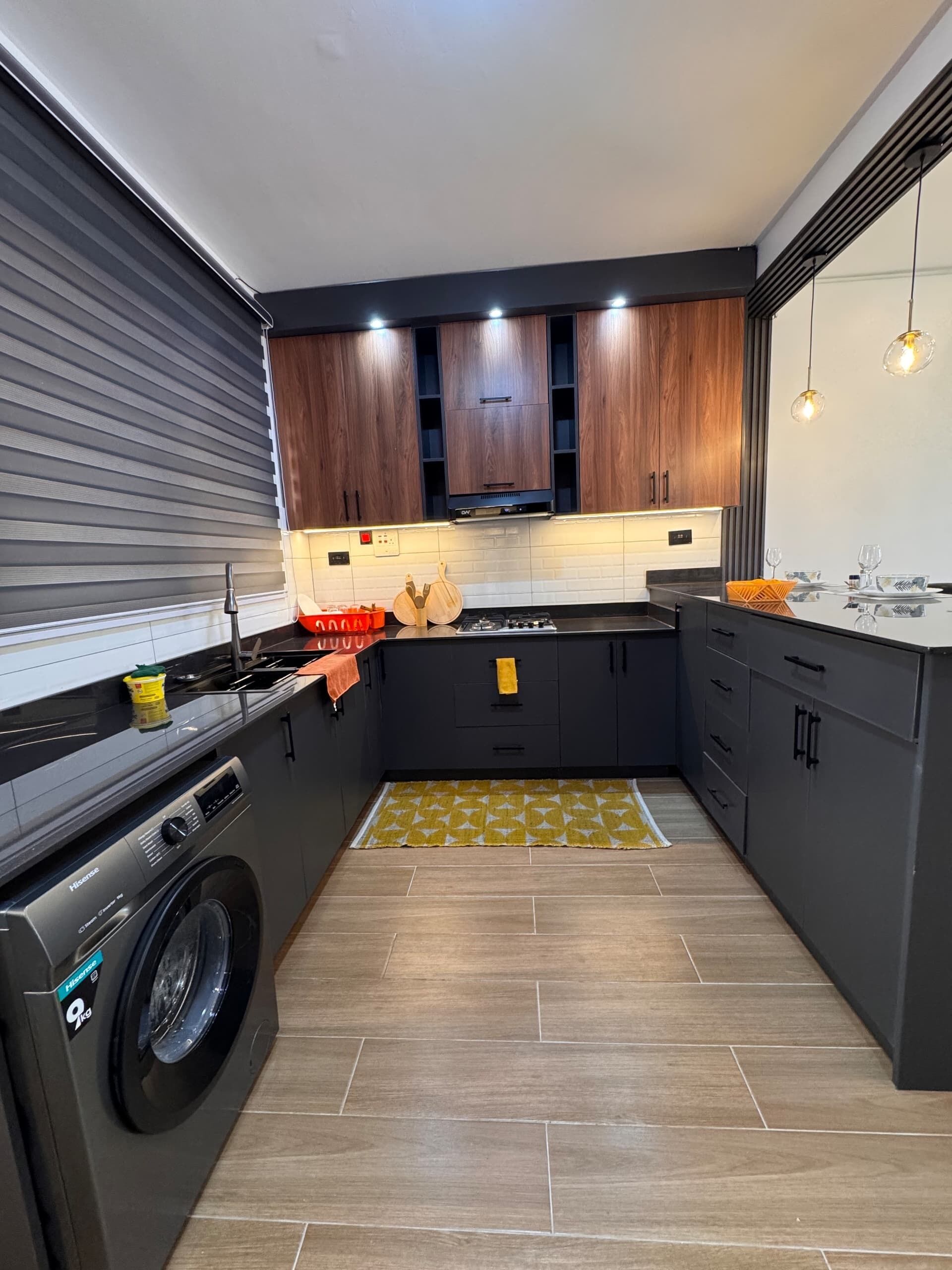 Una cocina moderna con armarios oscuros, lavadora integrada y una isla central con un tapete amarillo estampado.