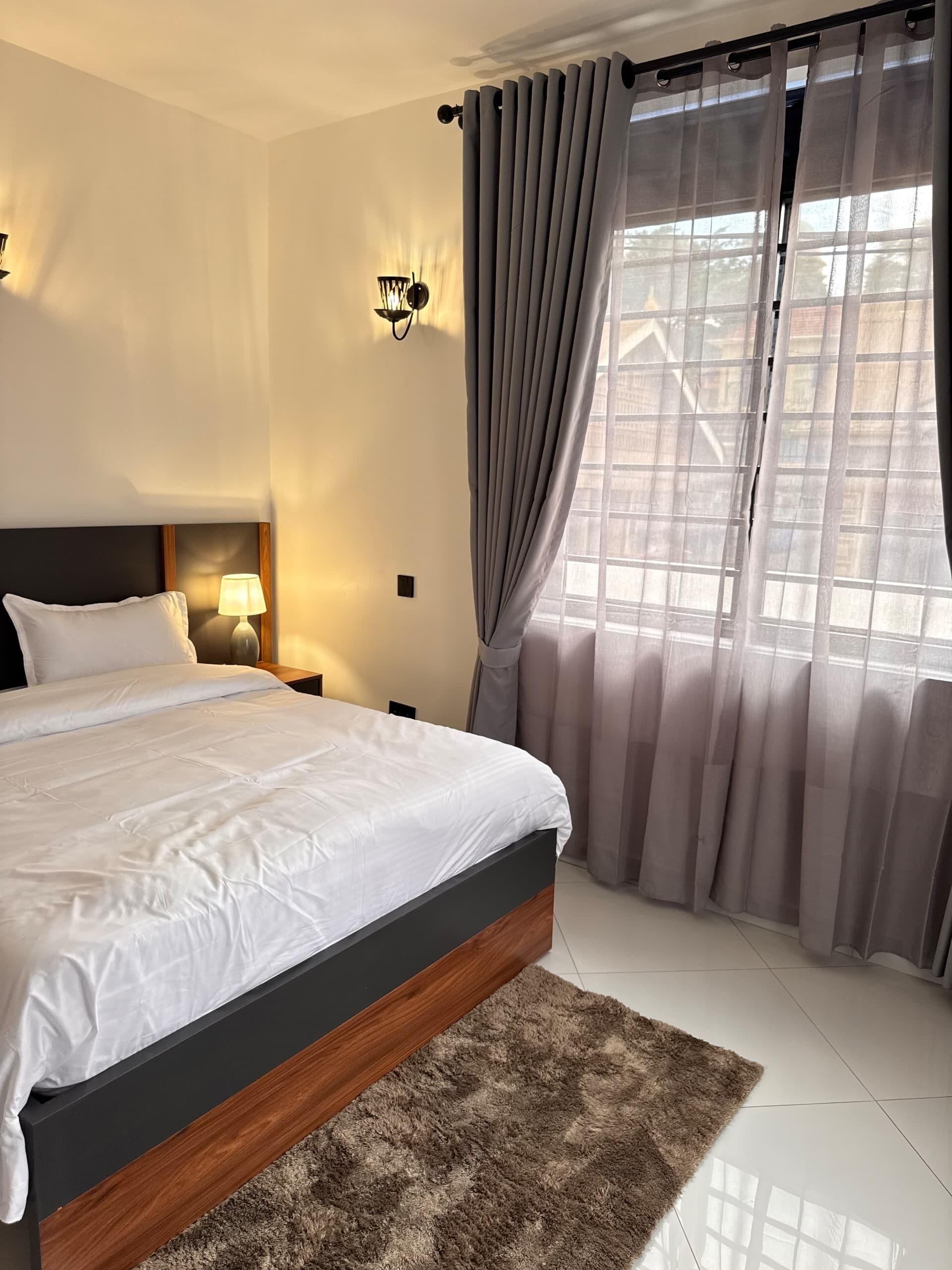 A bedroom with a bed, nightstands, a lamp, curtains, and a view of buildings through the window.
