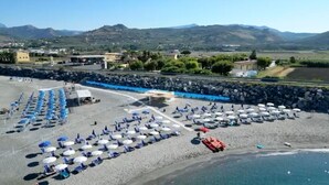 Ein Strand mit Reihen von Sonnenschirmen und Liegestühlen, einer felsigen Küste und Bergen im Hintergrund.