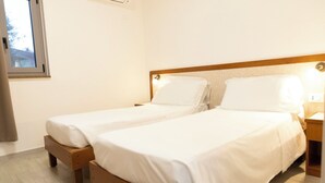 Una camera d'albergo con due letti singoli, una testiera in legno, un comodino con una lampada e una finestra che dà sulle parti esterne.
