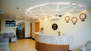 A hotel reception area with a curved reception desk, a flat-screen TV, a wall-mounted air conditioner, and clocks indicating different time zones.