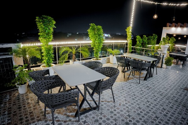 An outdoor seating area with tables and chairs, surrounded by potted plants and decorative lights.