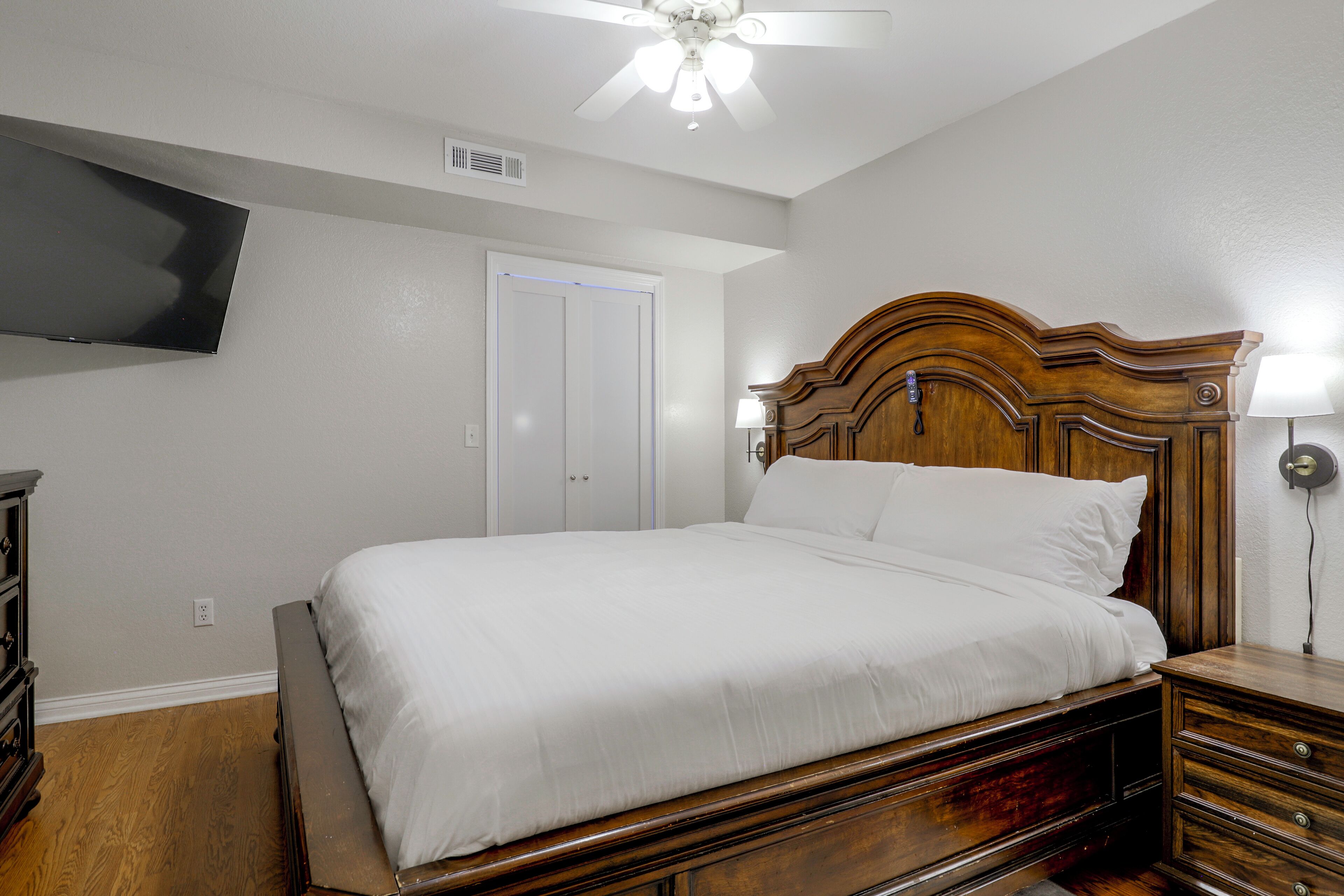 A bedroom with a wooden bed, a flat-screen TV mounted on the wall, a ceiling fan, and a bedside lamp.