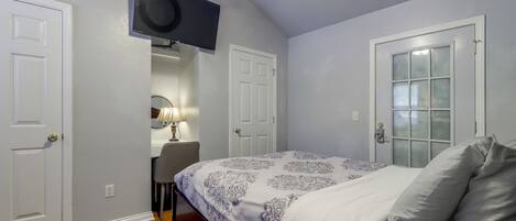 A bedroom with a bed, a desk with a chair, a television mounted on the wall, and a door with a window.