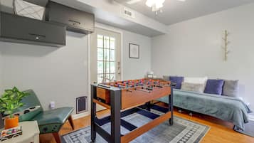 Una habitación con mesa de futbolín, sofá, sillón, pequeña mesa con una planta y ventilador de techo.