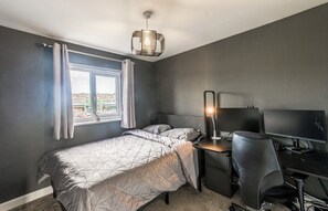 Ein Schlafzimmer mit einem Bett, einem Schreibtisch mit Computer, einem Stuhl und einem Fenster mit Vorhängen.