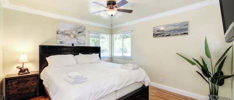 A bedroom with a bed, nightstand, lamp, ceiling fan, artwork, and a potted plant.