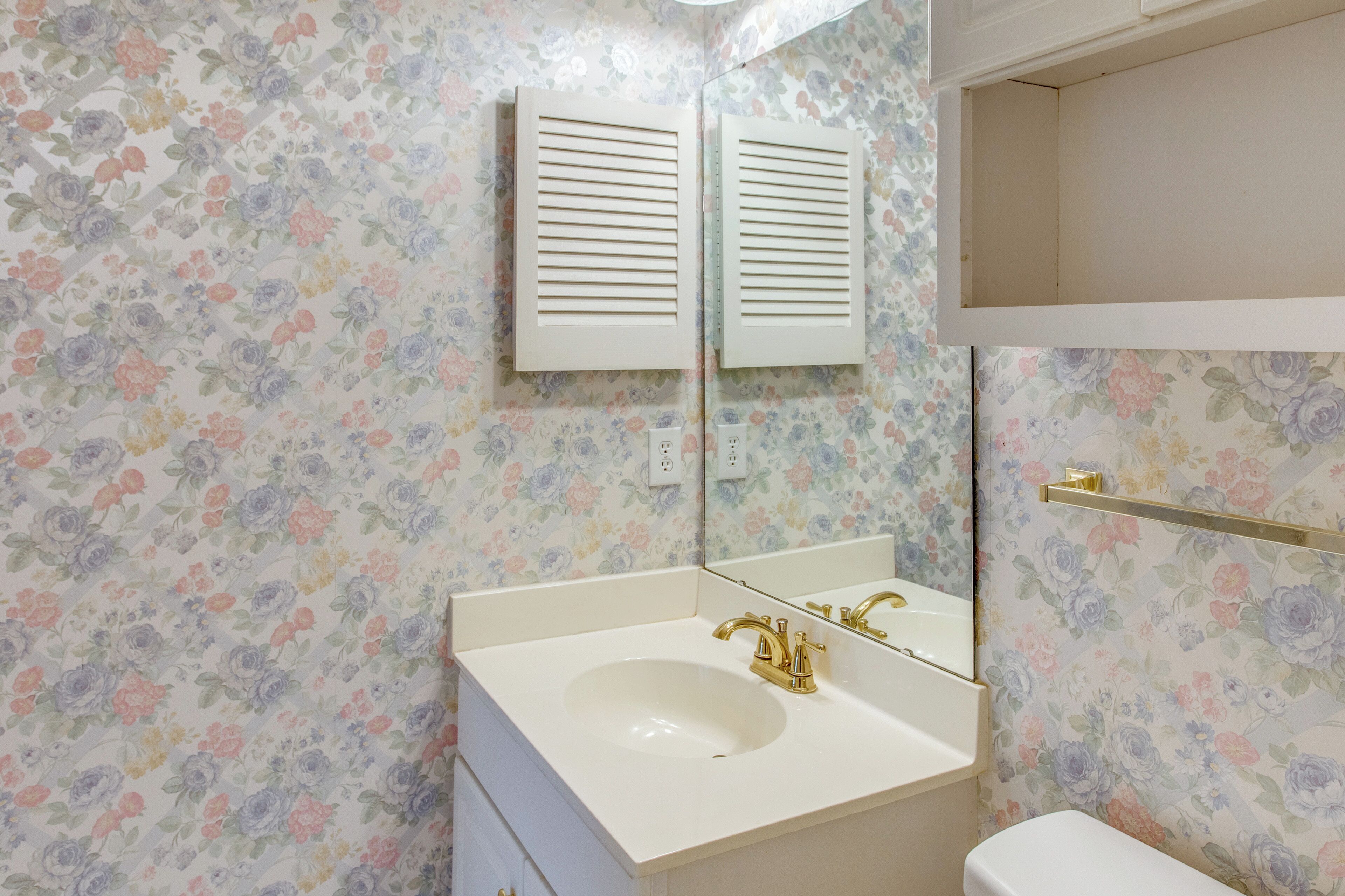 A bathroom with a floral wallpaper, a white sink with a gold faucet, and a mirror above the sink.