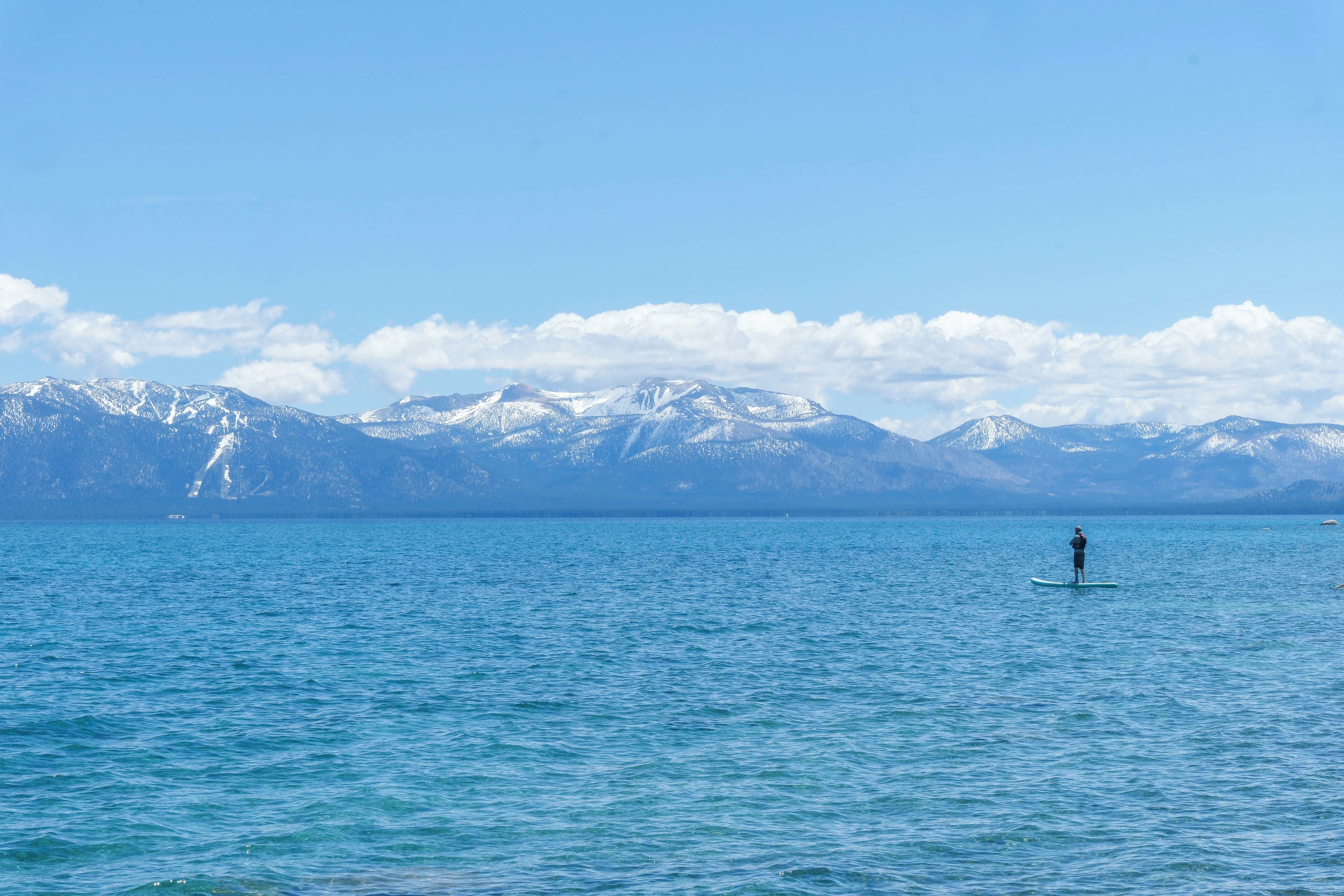 Eine Person steht auf einem Stand-Up-Paddleboard auf einem ruhigen See, im Hintergrund erheben sich schneebedeckte Berge.