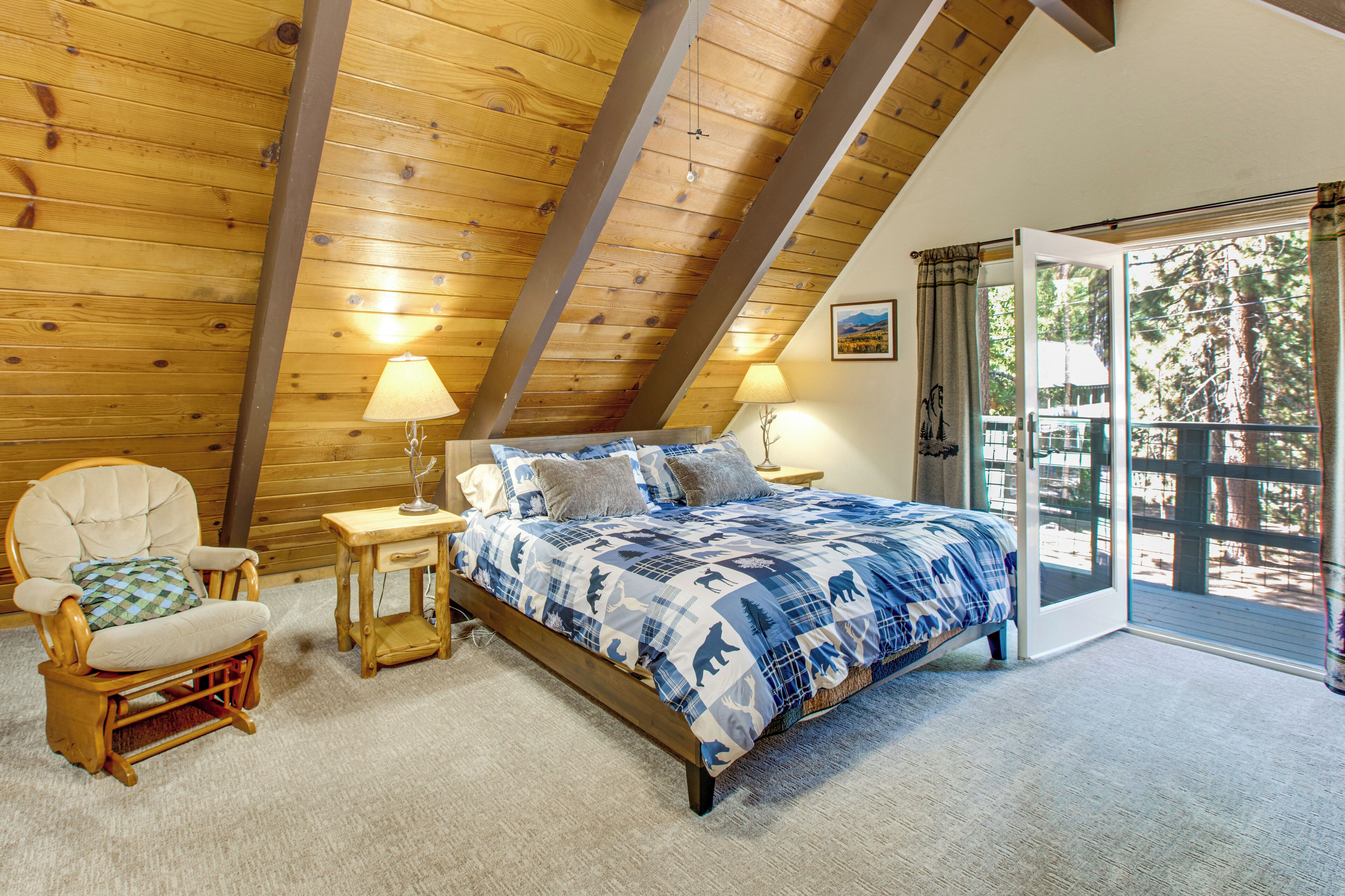Ein Schlafzimmer mit schrägen Wänden, einem Bett mit blauen und weißen Kissen, einem Holzstuhl, einem kleinen Nachttisch mit Lampe sowie Blick ins Freie durch eine Glastür.