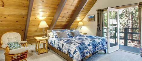 Ein Schlafzimmer mit schrägen Wänden, einem Bett mit blauen und weißen Kissen, einem Holzstuhl, einem kleinen Nachttisch mit Lampe sowie Blick ins Freie durch eine Glastür.