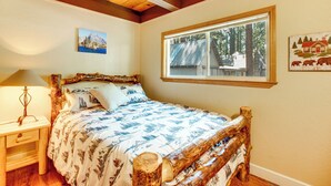 A bedroom with a wooden bed, a nightstand, a lamp, a window with a view of trees, and two wall art pieces. - Minutes to Slopes & Shore! Cozy Cabin w/ hot tub (South Lake Tahoe)