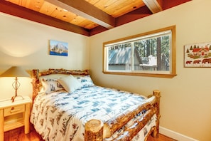 A bedroom with a wooden bed, a nightstand, a lamp, a window with a view of trees, and two wall art pieces. - Minutes to Slopes & Shore! Cozy Cabin w/ hot tub (South Lake Tahoe)