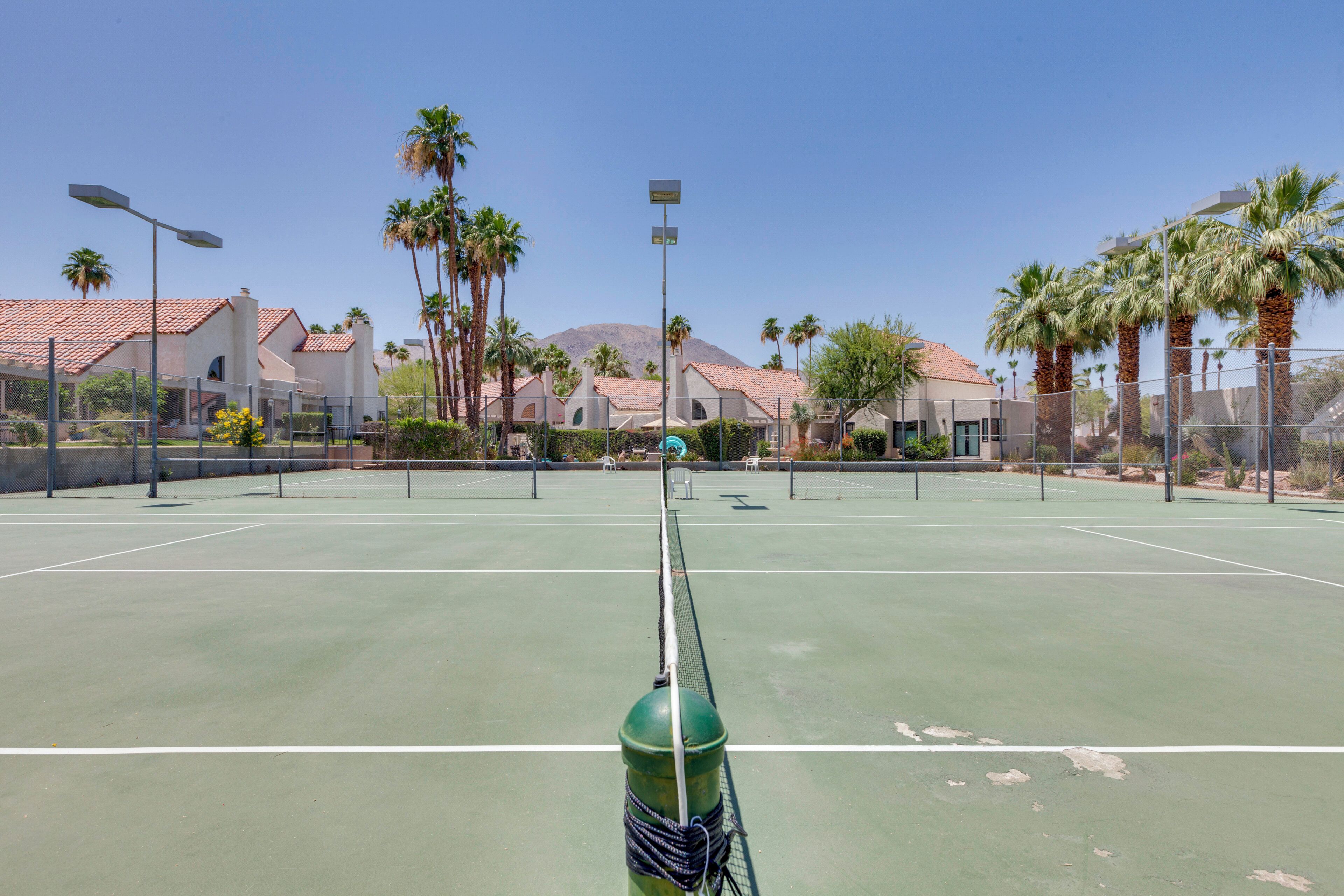 Una cancha de tenis con red, rodeada de palmeras y edificios con tejados rojos.