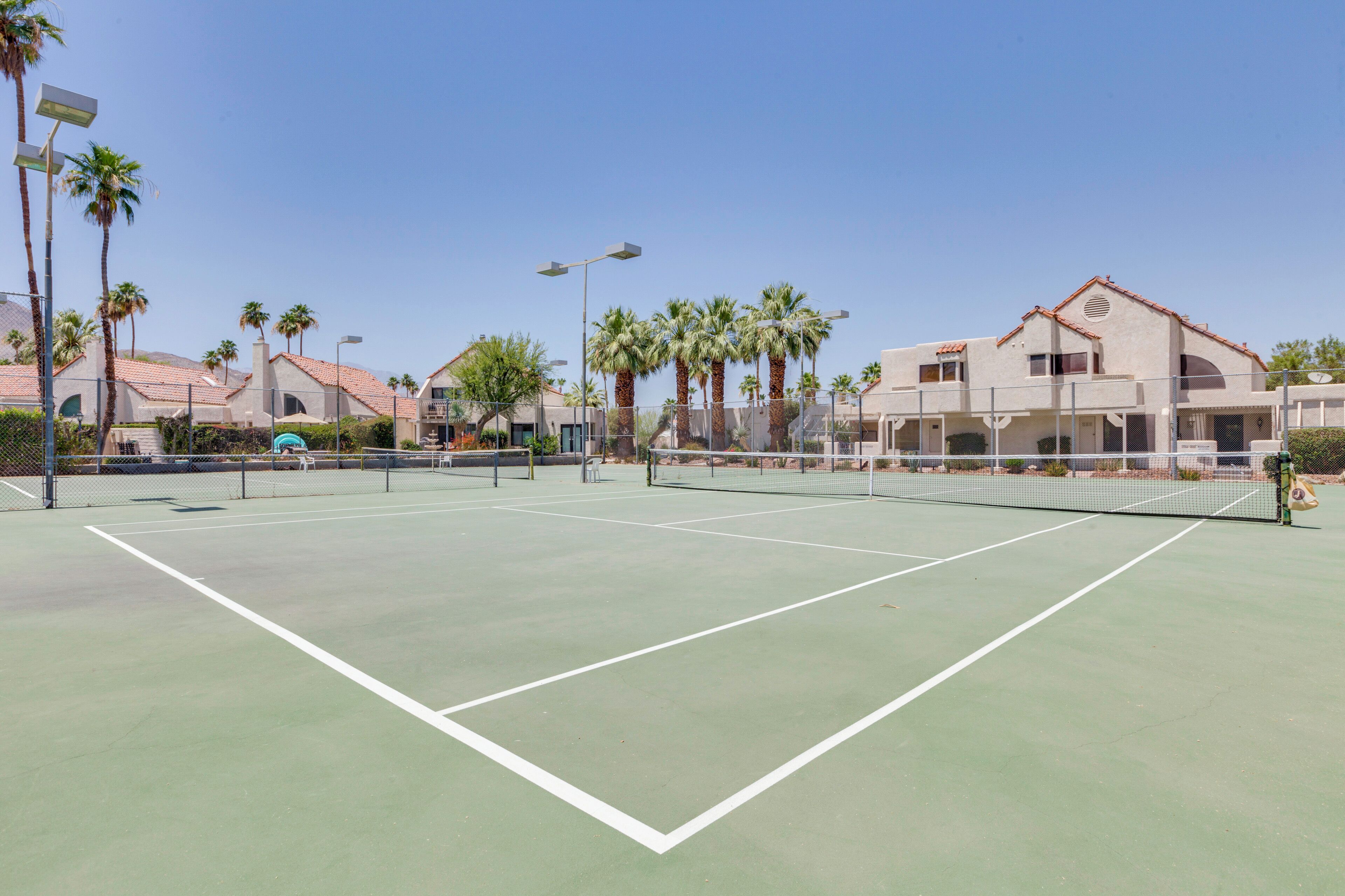A tennis court with a net, surrounded by palm trees and buildings with red-tiled roofs.