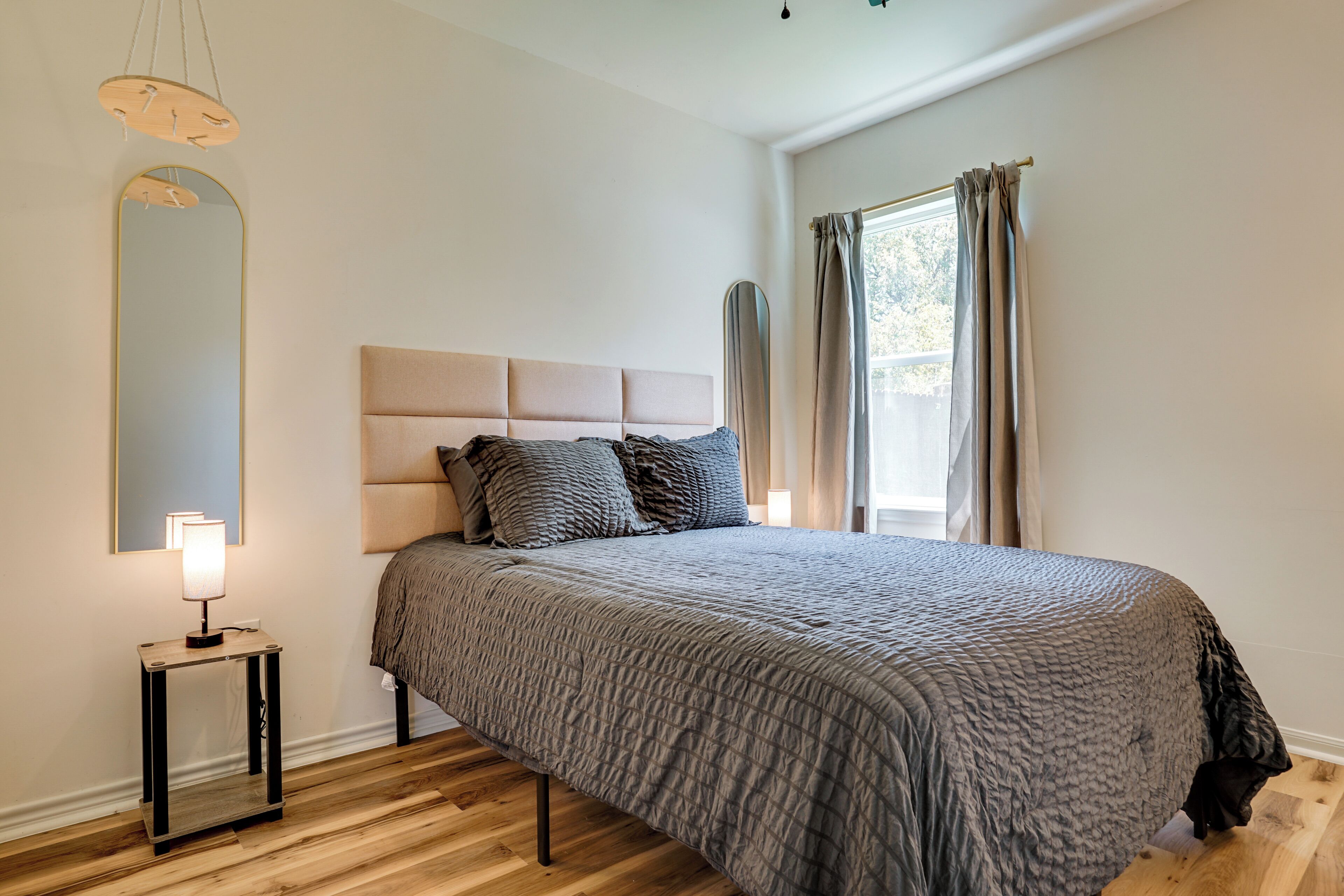 Ein Schlafzimmer mit einem großen Bett, einem Nachttisch mit Lampe, einem Spiegel und einem Fenster mit Vorhängen.