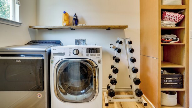 A laundry area with a front-loading washer and dryer, a shelf with cleaning supplies, and a storage unit with baskets. - Historic Southside Fort Worth Home w/ Fenced Yard! (Fort Worth)