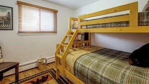 Uma cama de beliche com estrutura de madeira, uma pequena estante, uma janela com persianas e um quadro pendurado na parede.