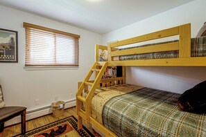 Ein Stockbett mit Holzrahmen, ein kleines Bücherregal, ein Fenster mit Jalousien und ein gerahmtes Bild an der Wand.