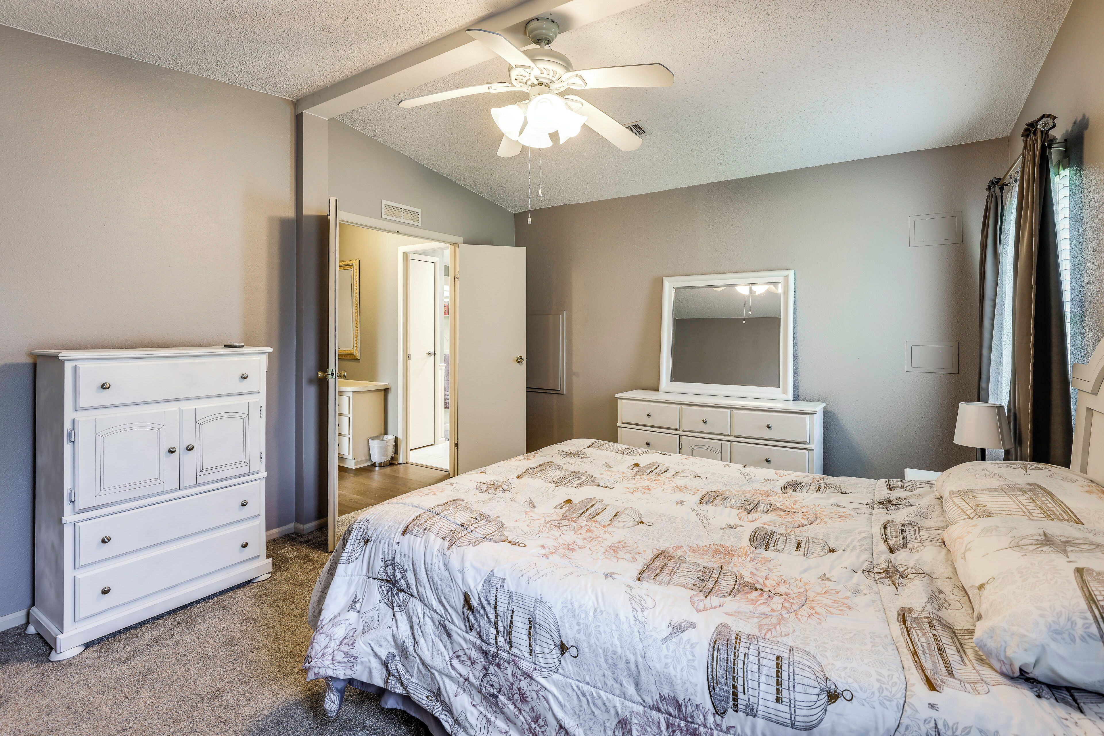 A bedroom with a bed, dresser, mirror, ceiling fan, and a door leading to another room.