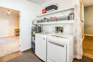 A compact kitchen with a refrigerator, a washing machine, and a water heater. There is a small shelf with storage baskets and a door leading to another room. - Yard & Screened Porch! Villages Escape (Lady Lake)