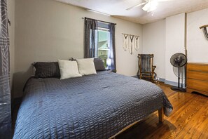 Un dormitorio con una cama grande, un sillón mecedor, un ventilador de pie y una ventana con cortinas.