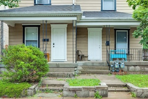 A residential building with a front porch, two doors, and a small garden area.