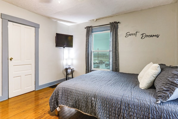A bedroom with a bed, a TV mounted on the wall, a window with curtains, and a door.