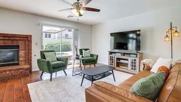 Een woonkamer met een open haard, groene fauteuils, een bruine leren bank, een flatscreen televisie en een plafondventilator.